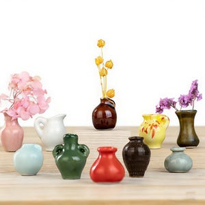 Könnte beinhalten: Eine Sammlung kleiner Keramikvasen in verschiedenen Farben, darunter Rosa, Weiß, Grün, Rot und Braun. Einige Vasen enthalten Trockenblumen, während andere leer sind. Die Vasen sind auf einer Holzoberfläche angeordnet.