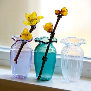 May include: Three small glass vases in shades of purple, teal, and clear, each holding a branch with yellow flowers. The vases have a ruffled top edge and are displayed on a white windowsill. The flowers add a pop of colour.