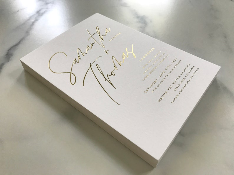 May include: A white wedding invitation with gold foil lettering. The invitation reads "Samantha + Thomas" and includes the wedding date, time, and location. The invitation is printed on thick, high-quality paper.