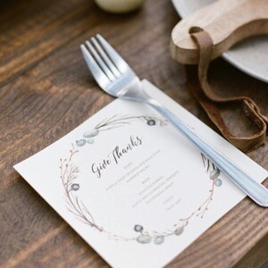 K&ouml;nnte beinhalten: Eine wei&szlig;e Karte mit einem Aquarell-Kranz-Design und dem Text "Give Thanks". Die Karte listet ein Men&uuml; f&uuml;r ein Thanksgiving-Essen auf, darunter lila S&uuml;&szlig;kartoffel- und Gr&uuml;nkohlsuppe, gebratenen Truthahn und Butternussk&uuml;rbissprossen. Eine silberne Gabel liegt auf der Karte.