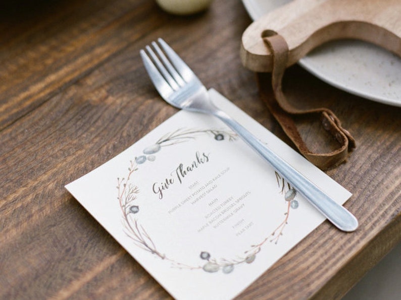 K&ouml;nnte beinhalten: Eine wei&szlig;e Karte mit einem Aquarell-Kranz-Design und dem Text "Give Thanks". Die Karte listet ein Men&uuml; f&uuml;r ein Thanksgiving-Essen auf, darunter lila S&uuml;&szlig;kartoffel- und Gr&uuml;nkohlsuppe, gebratenen Truthahn und Butternussk&uuml;rbissprossen. Eine silberne Gabel liegt auf der Karte.