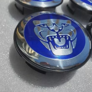 May include: A set of Jaguar car wheel center caps. Each cap features a silver Jaguar emblem on a blue background, surrounded by a chrome-like finish. The caps are circular and appear to be new.