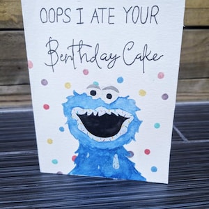 May include: A greeting card with the text "OOPS I ATE YOUR Birthday Cake" above a blue monster illustration. The card has colorful polka dots and is on a dark surface.