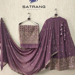 May include: A three-piece ensemble in a dusty rose hue, featuring intricate gold embroidery and embellishments. The set includes a top, a draped shawl, and a skirt, all adorned with delicate details. The "SATRANG BOUTIQUE" logo is visible at the top.