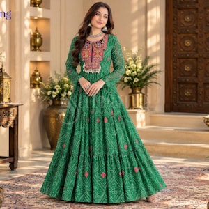 May include: A floor-length green dress with long sleeves and a tiered skirt. The fabric features a white geometric pattern and small red accents. The bodice has a prominent rectangular panel with intricate multi-colored embroidery and tassels.