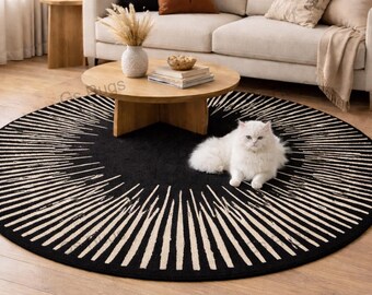May include: A round black rug with a white sunburst design. A white cat is lying on the rug in front of a beige sofa. The rug is on a hardwood floor in a living room setting.