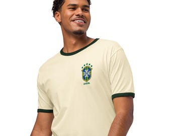 Könnte beinhalten: Ein cremefarbenes Ringer-T-Shirt mit dunkelgrünen Abschlüssen an Kragen und Ärmeln. Auf der Brust befindet sich ein kleines gesticktes Emblem der brasilianischen Fußballnationalmannschaft mit dem CBF-Logo, fünf grünen Sternen und dem Wort BRASIL.