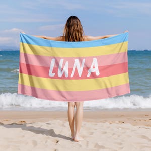 May include: A beach towel with horizontal stripes in shades of blue, yellow, and pink. The word "LUNA" is printed in large white letters across the center of the towel. The towel is held up on a beach with the ocean in the background.