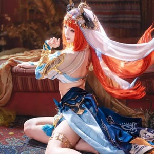 Puede incluir: Un disfraz de cosplay que incluye un top corto blanco y azul con detalles dorados, una falda azul a juego con patrones blancos intrincados y un tocado con cuernos y un velo blanco fluido. El conjunto se completa con un largo cabello naranja vibrante.