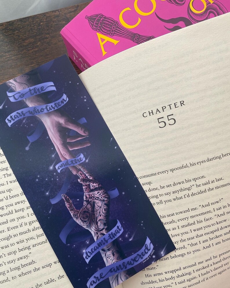 May include: A bookmark with a dark blue background and a starry sky. Two hands are reaching towards each other with the text "To the stars who listen" and "dreams that are answered" on the bookmark.