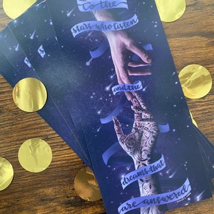 May include: A stack of blue and purple bookmarks with a quote that reads "To the stars who listen and the dreams that are answered." The quote is written in a white cursive font. The bookmarks feature a hand reaching out to another hand with a starry sky background.