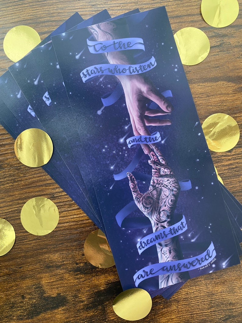 May include: A stack of blue and purple bookmarks with a quote that reads "To the stars who listen and the dreams that are answered." The quote is written in a white cursive font. The bookmarks feature a hand reaching out to another hand with a starry sky background.