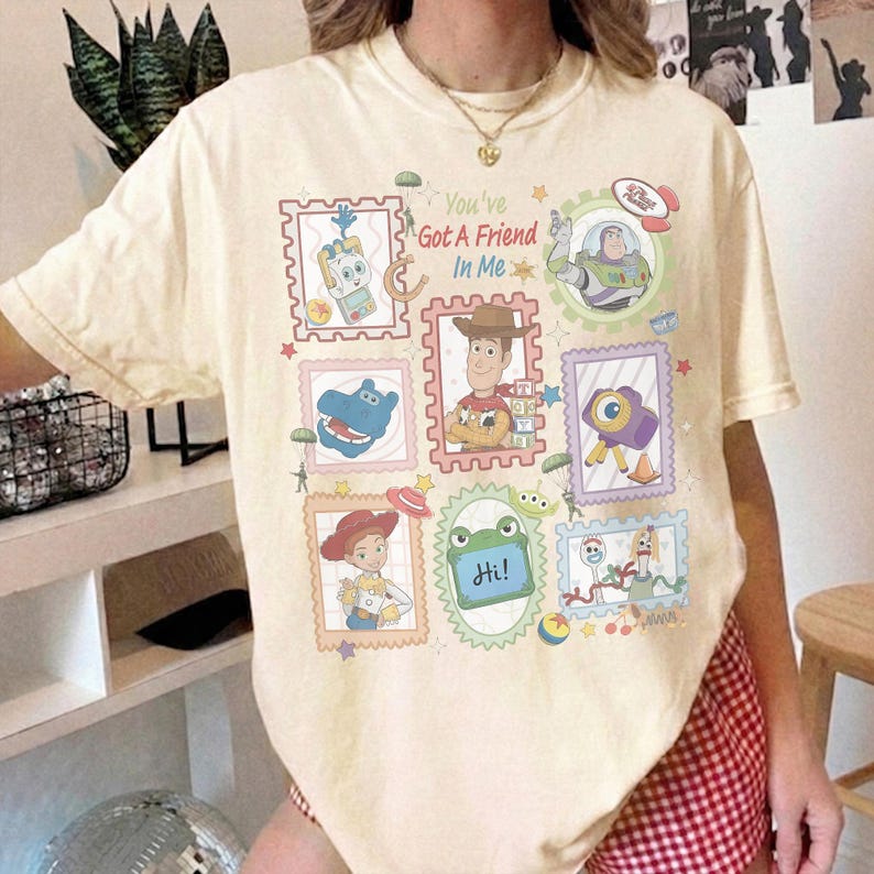 Puede incluir: Una camiseta de color crema con un estampado gr&aacute;fico de varios personajes de Toy Story dispuestos en marcos con forma de sello postal. El texto "You've Got A Friend In Me" est&aacute; impreso en el centro, rodeado de ilustraciones de Woody, Buzz Lightyear, Jessie y otras figuras ic&oacute;nicas de la serie de pel&iacute;culas.