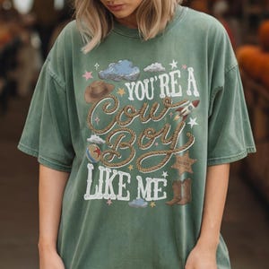 May include: A sage green short-sleeve t-shirt featuring a graphic print with the text "YOU'RE A COW BOY LIKE ME" written in white rope-style lettering. The design includes a brown cowboy hat, a rocket, a sheriff badge, and cowboy boots.