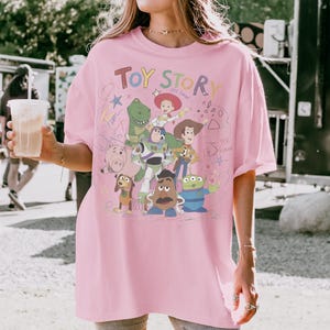 Puede incluir: Una camiseta rosa claro de gran tama&ntilde;o con un gr&aacute;fico colorido de personajes de la pel&iacute;cula animada "Toy Story". El dise&ntilde;o incluye a Woody, Buzz Lightyear, Rex y otros personajes, con las palabras "Toy Story" en una fuente l&uacute;dica.