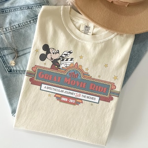 May include: Cream-coloured t-shirt featuring Mickey Mouse holding a film clapper. The shirt has a retro design with the text "The Great Movie Ride" and "A Spectacular Journey Into The Movies 1989-2017". The shirt is folded with a hat and jeans.