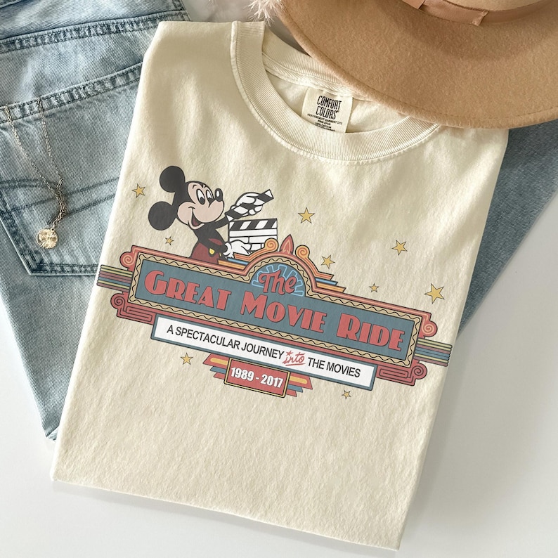 May include: Cream-coloured t-shirt featuring Mickey Mouse holding a film clapper. The shirt has a retro design with the text "The Great Movie Ride" and "A Spectacular Journey Into The Movies 1989-2017". The shirt is folded with a hat and jeans.