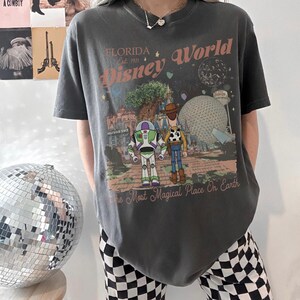 May include: Dark gray t-shirt featuring a graphic of Disney World in Florida. The design includes Buzz Lightyear, Woody, and the Hollywood Tower Hotel. Text reads "Florida Disney World Est. 1971" and "The Most Magical Place On Earth."