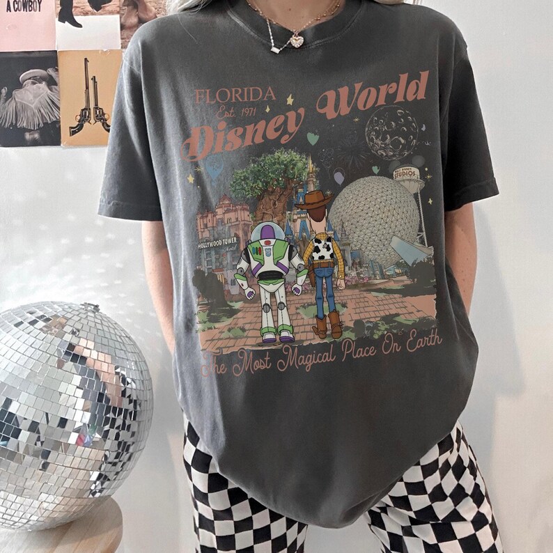 May include: Dark gray t-shirt featuring a graphic of Disney World in Florida. The design includes Buzz Lightyear, Woody, and the Hollywood Tower Hotel. Text reads "Florida Disney World Est. 1971" and "The Most Magical Place On Earth."