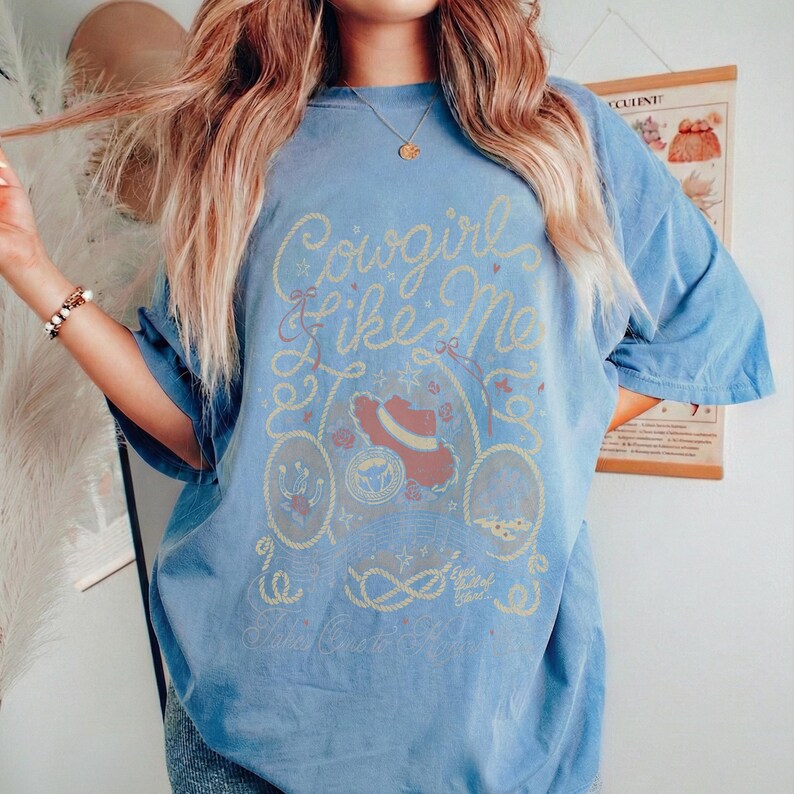 May include: A blue short-sleeve t-shirt featuring a graphic print in yellow and red tones. The design includes the text Cowgirl Like Me, a cowboy hat, a horseshoe, and rope motifs. The shirt has a crew neckline and a relaxed, loose fit.