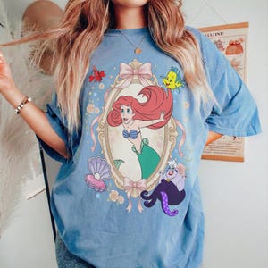 Puede incluir: Camiseta azul claro con una ilustraci&oacute;n enmarcada de Ariel de La Sirenita, con Flounder, Sebasti&aacute;n y &Uacute;rsula. El dise&ntilde;o incluye lazos rosas, flores y una concha. La camiseta tiene mangas cortas y un corte holgado.