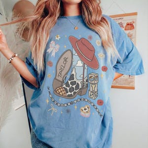 May include: A blue short-sleeve t-shirt featuring a graphic print of a cow-print cowboy boot, a red cowboy hat, and blocks with the letters T, S, and the number 13. The word TAYLOR is printed on the boot, surrounded by small flowers and stars.