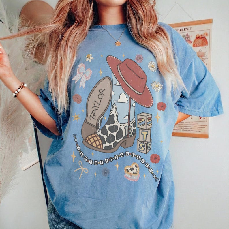 May include: A blue short-sleeve t-shirt featuring a graphic print of a cow-print cowboy boot, a red cowboy hat, and blocks with the letters T, S, and the number 13. The word TAYLOR is printed on the boot, surrounded by small flowers and stars.