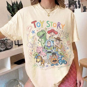 May include: A cream-coloured short-sleeve t-shirt featuring a colourful graphic print of Toy Story characters including Woody, Buzz Lightyear, Jessie, Rex, Hamm, Slinky Dog, Mr. Potato Head, and Aliens, surrounded by scribbled drawings and text.