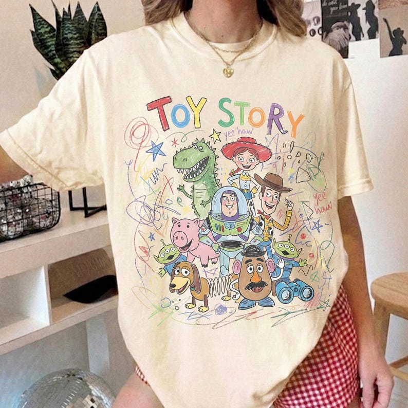 May include: A cream-coloured short-sleeve t-shirt featuring a colourful graphic print of Toy Story characters including Woody, Buzz Lightyear, Jessie, Rex, Hamm, Slinky Dog, Mr. Potato Head, and Aliens, surrounded by scribbled drawings and text.