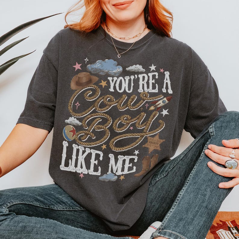 May include: A charcoal gray short-sleeve t-shirt featuring a graphic print with the text "YOU'RE A COWBOY LIKE ME" in white lettering. The design includes a brown cowboy hat, a lasso, a rocket, a sheriff star, and small blue clouds and stars.