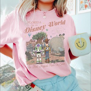 May include: Pink t-shirt with a graphic of Disney World landmarks and characters, including Buzz Lightyear and Woody. The shirt has the text "Florida Disney World Est. 1971" and "The Most Magical Place On Earth". A white mug with a yellow smiley face is held.