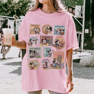 May include: A light pink t-shirt featuring Mickey Mouse in various travel-themed designs. The shirt includes images of Mickey Mouse in front of landmarks from Mexico, London, Norway, Japan, China, Morocco, Germany, USA, Italy, Canada, and France.
