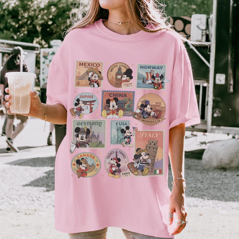 May include: A light pink t-shirt featuring Mickey Mouse in various travel-themed designs. The shirt includes images of Mickey Mouse in front of landmarks from Mexico, London, Norway, Japan, China, Morocco, Germany, USA, Italy, Canada, and France.