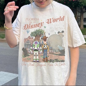 May include: Cream-colored t-shirt featuring a Disney World graphic. The design includes the text "Florida Est. 1971 Disney World" and illustrations of Buzz Lightyear, Woody, and various park landmarks. The phrase "The Most Magical Place On Earth" is also present.