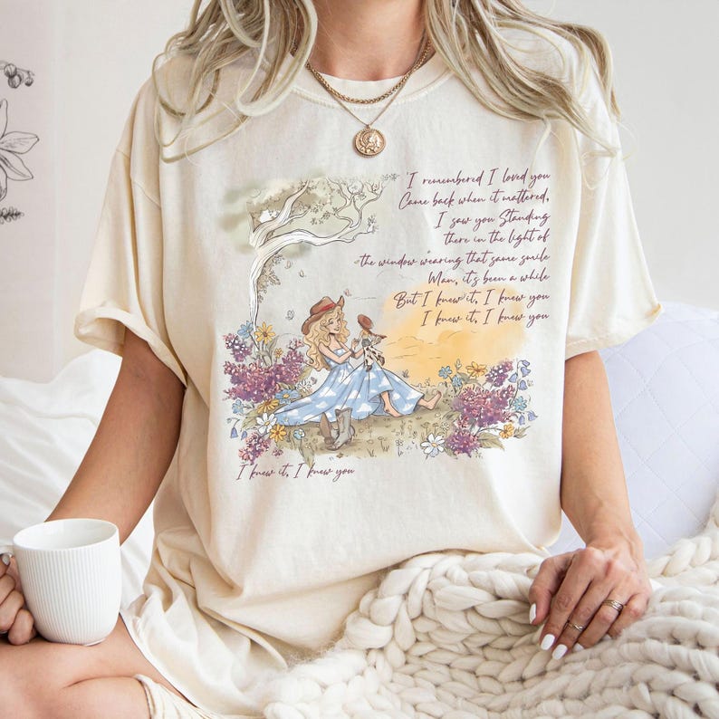 May include: A cream-colored short-sleeve t-shirt featuring a graphic illustration of a person in a blue dress and cowboy boots sitting in a field of flowers. The design includes printed lyrics and the text, I knew it, I knew you.