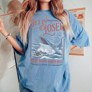 Puede incluir: Camiseta azul claro con el texto "GET IN LOSER" en rojo y azul. El gr&aacute;fico presenta una ballena en el oc&eacute;ano con una persona en un bote. Tambi&eacute;n se incluye el texto "WE'RE DOING GOD'S WILL" y "JONAH 1:17".