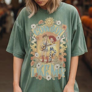 May include: A sage green short-sleeve t-shirt featuring a graphic print of two characters, one playing a guitar, surrounded by flowers and a sun. The text "COWBOY LIKE ME" is printed in light blue letters above and below the central illustration.