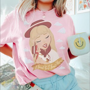 May include: A light pink t-shirt featuring a graphic illustration of a blonde person wearing a maroon hat and holding a small doll. The design includes white clouds and a tan banner with the text I Knew It, I Knew You printed in brown.
