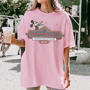 May include: Pale pink t-shirt with a graphic of Mickey Mouse holding a film clapperboard. The design includes the text "The Great Movie Ride" and "A Spectacular Journey Into The Movies 1989-2017" with star accents.