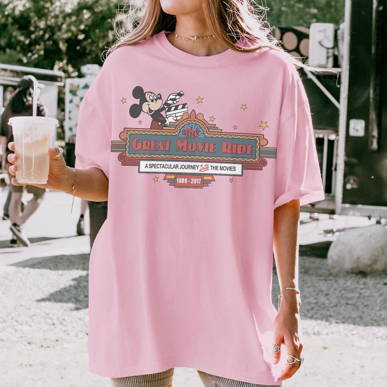 May include: Pale pink t-shirt with a graphic of Mickey Mouse holding a film clapperboard. The design includes the text "The Great Movie Ride" and "A Spectacular Journey Into The Movies 1989-2017" with star accents.