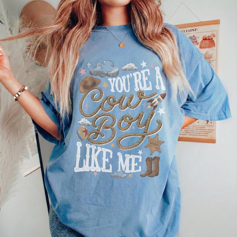 May include: A light blue short-sleeve t-shirt featuring a graphic print with the text "YOU'RE A COWBOY LIKE ME" in white lettering. The design includes a brown lasso, a cowboy hat, a rocket, a sheriff badge, and brown cowboy boots.