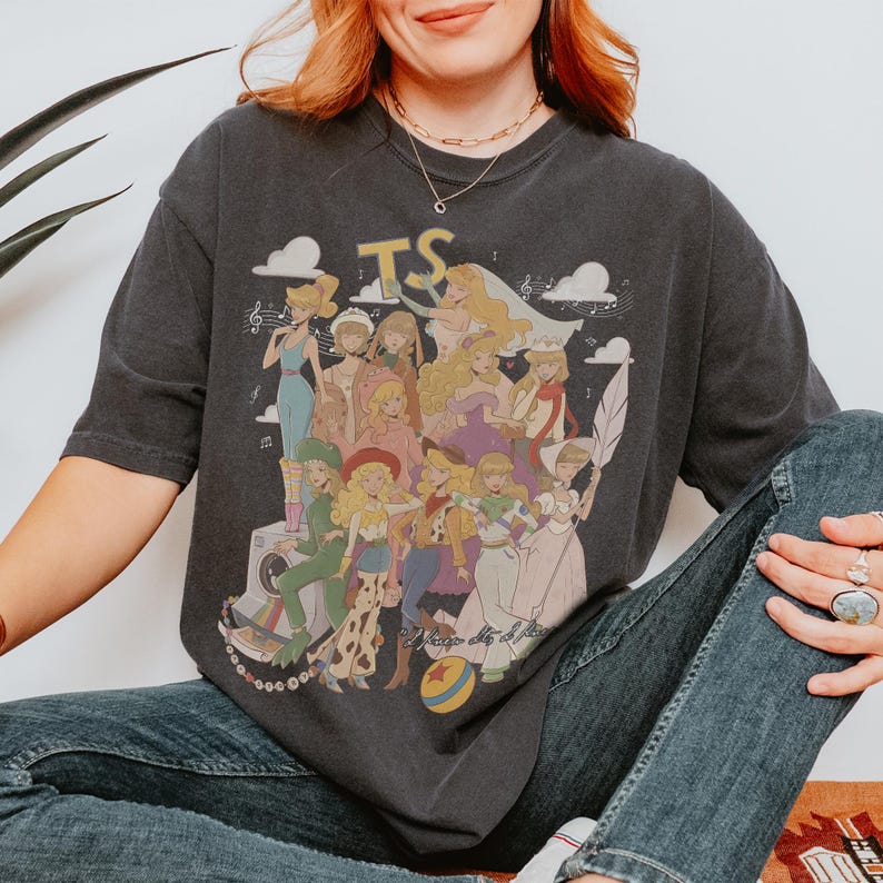 Puede incluir: Una camiseta gris carb&oacute;n de manga corta con un estampado gr&aacute;fico de varios personajes femeninos animados con diversos atuendos. El dise&ntilde;o incluye el texto TS en la parte superior, notas musicales, nubes, una c&aacute;mara y una pelota con estrellas.