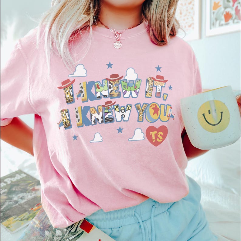 K&ouml;nnte beinhalten: Ein hellrosa T-Shirt mit einem Grafikdruck und dem Text I KNEW IT, I KNEW YOU. Die Buchstaben sind mit Mustern gef&uuml;llt, die von Cowboy- und Weltraum-Ranger-Figuren inspiriert sind, akzentuiert durch kleine braune H&uuml;te, blaue Sterne und ein rotes Herz.