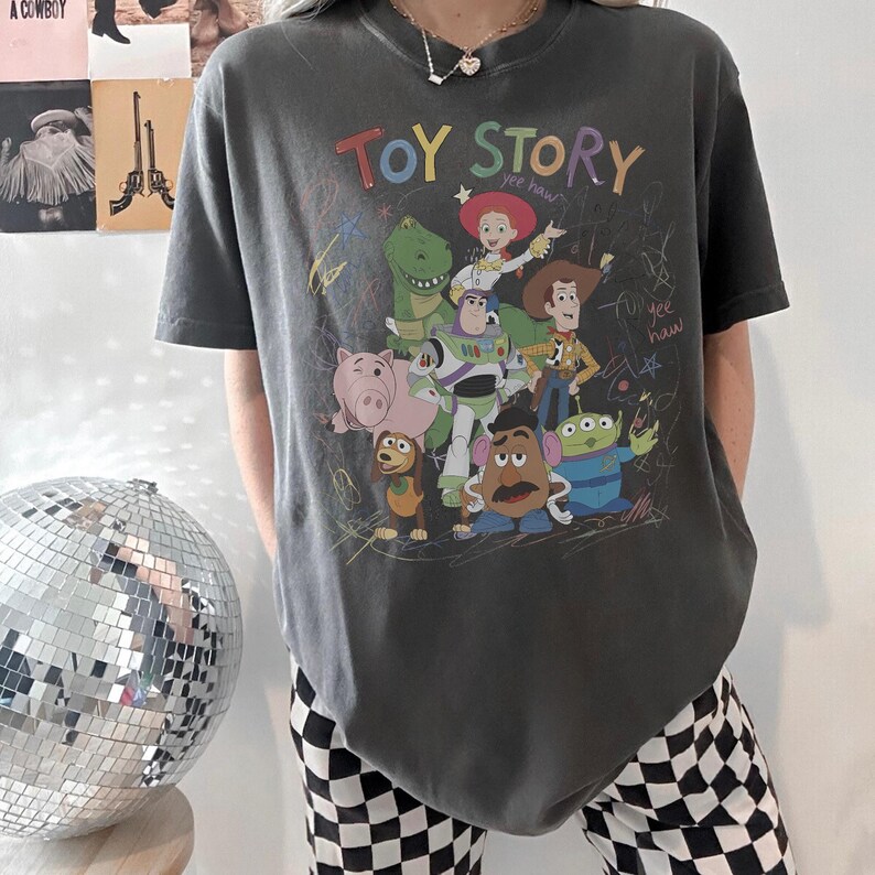 Puede incluir: Camiseta gris oscuro con las palabras "TOY STORY" en letras coloridas y un gr&aacute;fico de personajes de la pel&iacute;cula animada. El dise&ntilde;o incluye a Woody, Buzz Lightyear y otros personajes. La camiseta tiene mangas cortas.