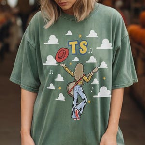 May include: A sage green t-shirt featuring a graphic of a person with long hair, wearing a yellow plaid shirt and cow-print bell-bottom pants, holding a guitar and a red hat. The design includes white clouds, yellow stars, musical notes, and the text TS.