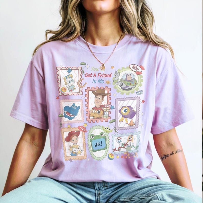 Puede incluir: Una camiseta de manga corta de color morado claro con un estampado gr&aacute;fico de varios personajes de dibujos animados de Toy Story dispuestos en marcos estilo sello postal. El texto "You Got A Friend In Me" aparece en el centro.