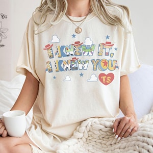 K&ouml;nnte beinhalten: Ein cremefarbenes Kurzarm-T-Shirt mit einem Grafikdruck, der den Text I KNEW IT, I KNEW YOU und TS in einem roten Herzen zeigt. Das Design besteht aus blauen Buchstaben, die mit Mustern von Cowboyh&uuml;ten und Kuhflecken gef&uuml;llt sind.