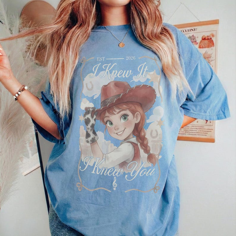 May include: A blue short-sleeve t-shirt featuring a graphic of a cartoon cowgirl wearing a brown hat and a cow-print glove. The text reads "I Knew It" at the top, "EST 2026" in the center, and "I Knew You" at the bottom, surrounded by clouds.