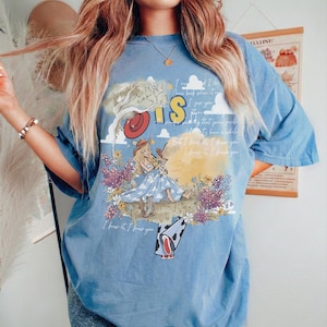 May include: A blue short-sleeve t-shirt featuring a graphic print of a character in a blue dress and cowboy boots sitting in a field of flowers. The design includes a red hat, clouds, and text that reads I knew it, I knew you.