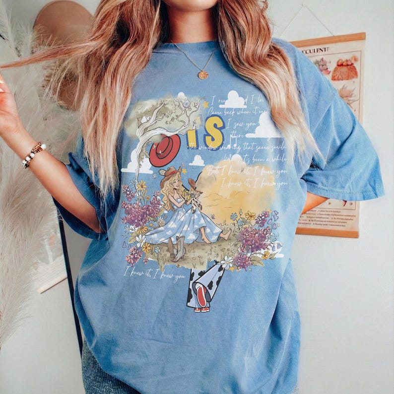 May include: A blue short-sleeve t-shirt featuring a graphic print of a character in a blue dress and cowboy boots sitting in a field of flowers. The design includes a red hat, clouds, and text that reads I knew it, I knew you.
